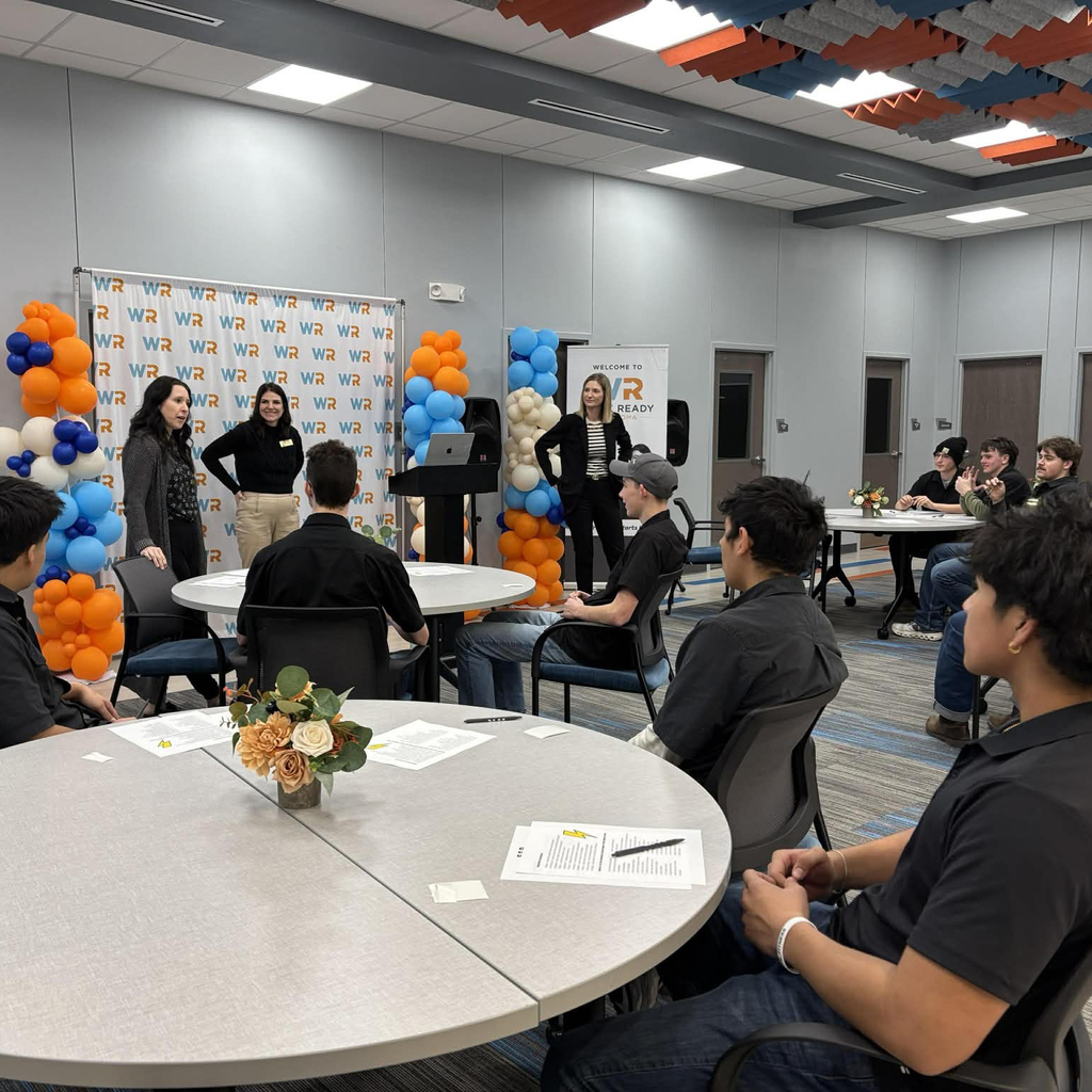 Several people in a classroom setting with presenters at the front of the room.