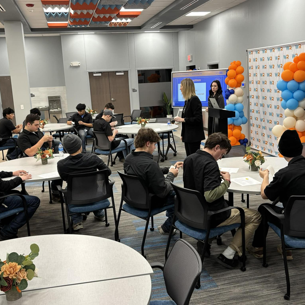 Several people in a classroom setting with presenters at the front of the room.