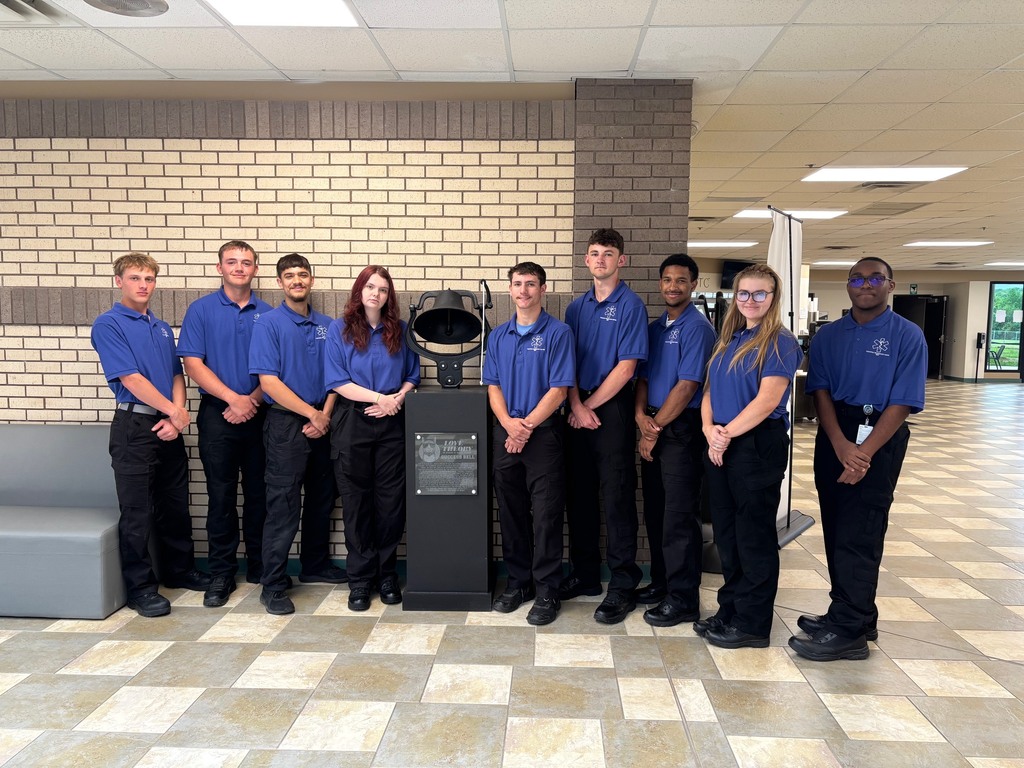 A group of 9 students wearing blue shirts standing next to the success bell.
