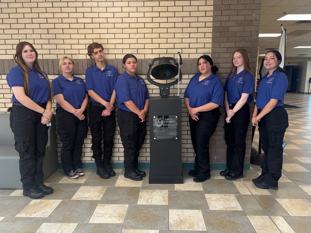 A group of 7 students wearing blue shirts standing next to the success bell.