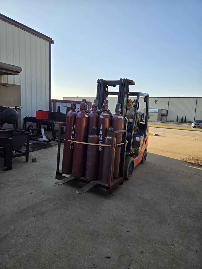 A student behind the wheel of a forklift transporting a rack of cylinders.