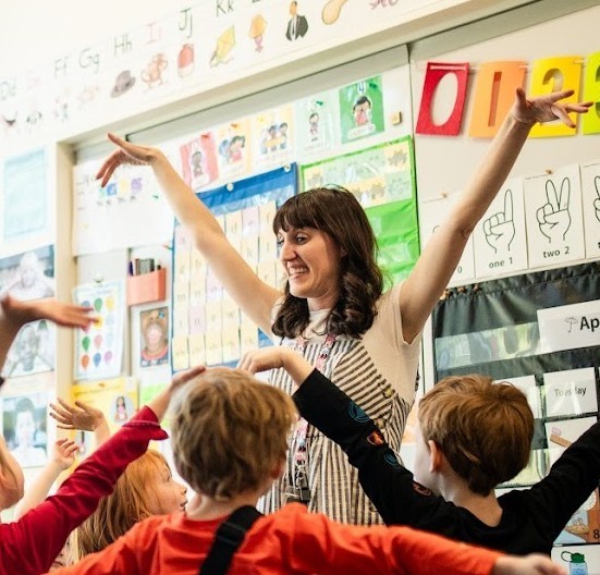 kindergarten teacher with arms up high showing students how high a sunflower grows a few student s backs can be seen