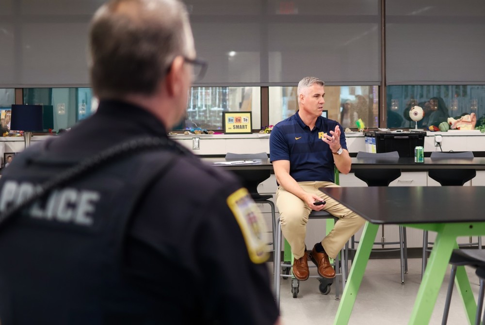 a man speaks inside a classroom while a police officer looks on
