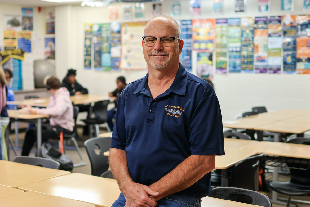 a man posing for a photo in a classroom with kids at desks