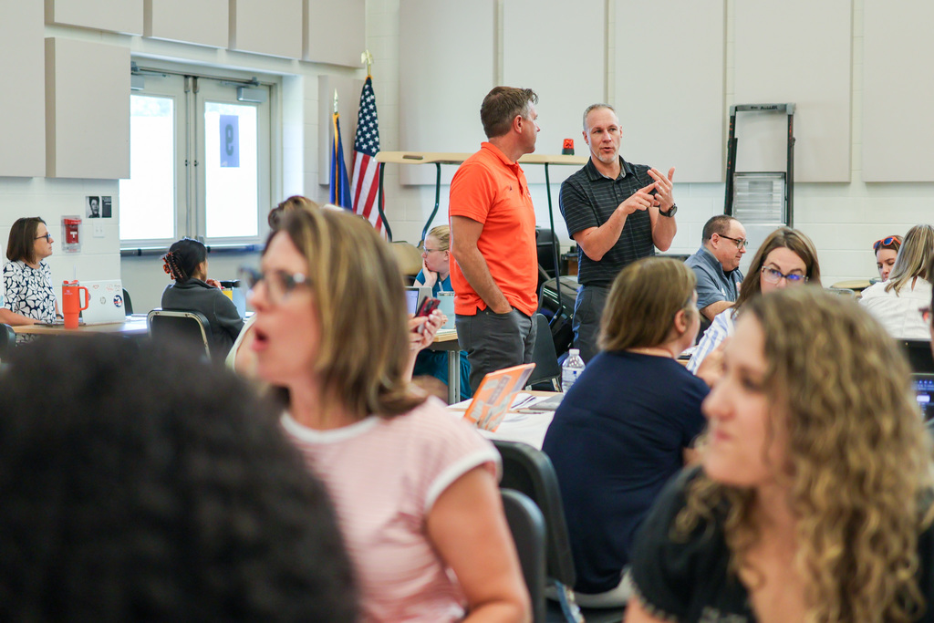two men stand and talk in a room full of people
