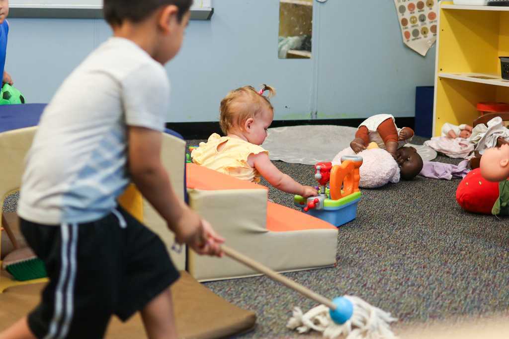 a baby plays with a toy while a young boy uses a toy mop inside a classroom