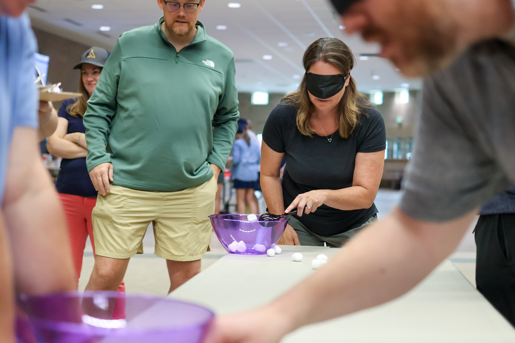 a blind folded woman tries to put cotton balls in a bowl with a spoon