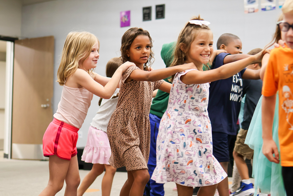 students with hands on each others shoulders walk in a train across a classroom