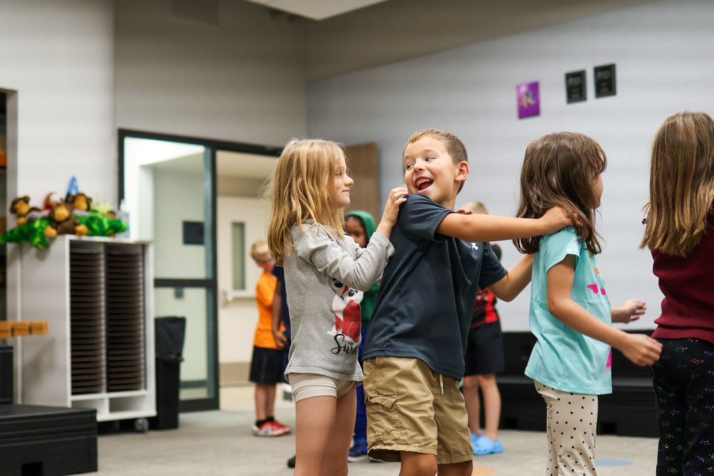 students with hands on each others shoulders walk in a train across a classroom