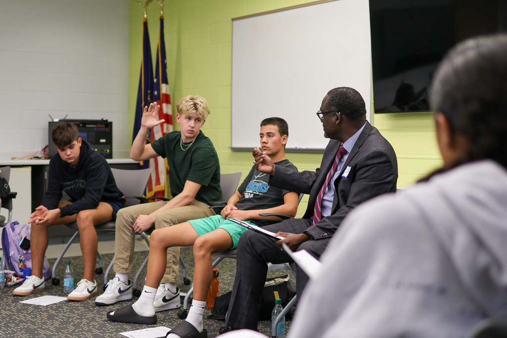 a boy holds his hand up sitting in a conference room with other students
