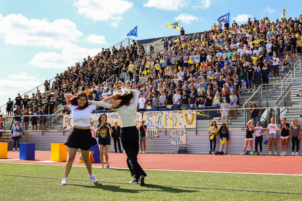 two girls perform a dance in front of others gathered in a football stadium
