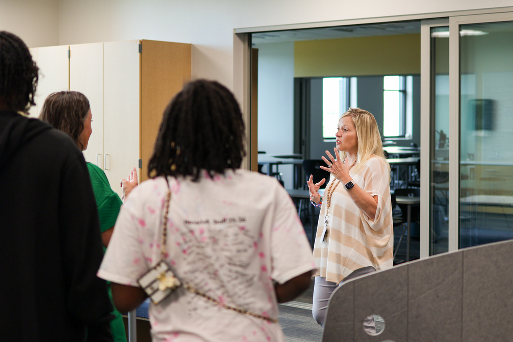 a woman talking to a group of people gathered inside a school classroom
