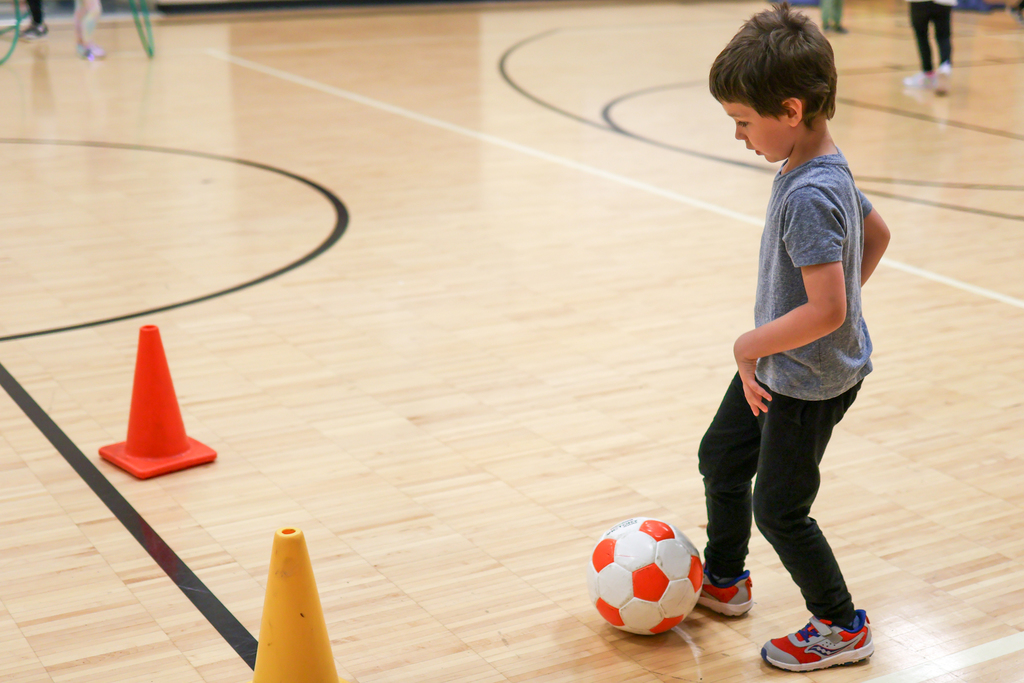 a boy kicking a soccer ball around cones in a gym