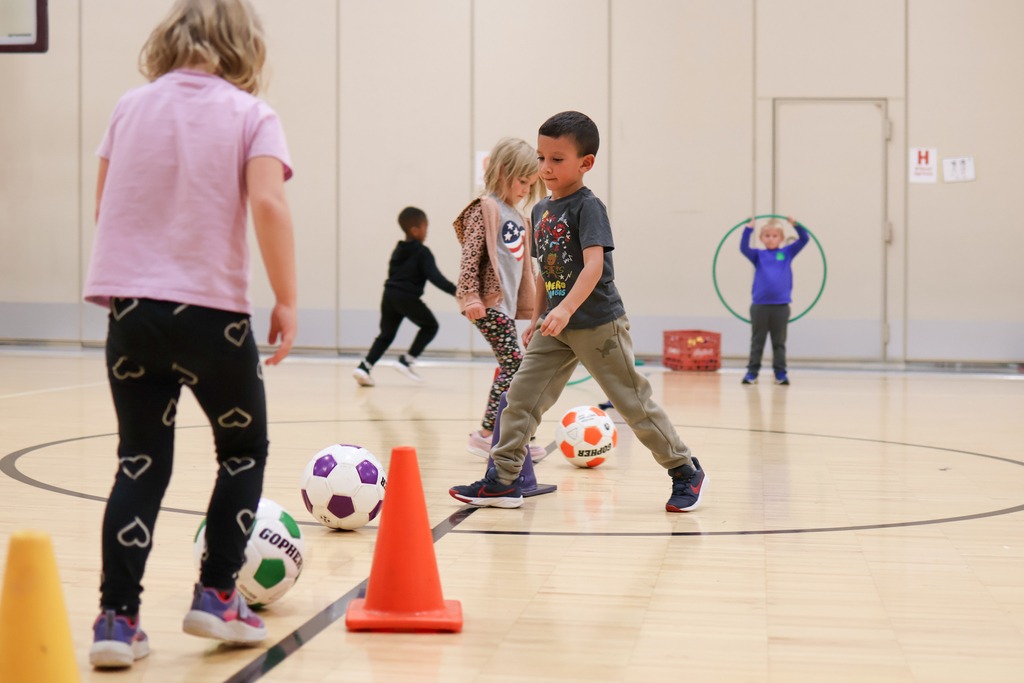 kids kicking soccer balls around cones inside a gym