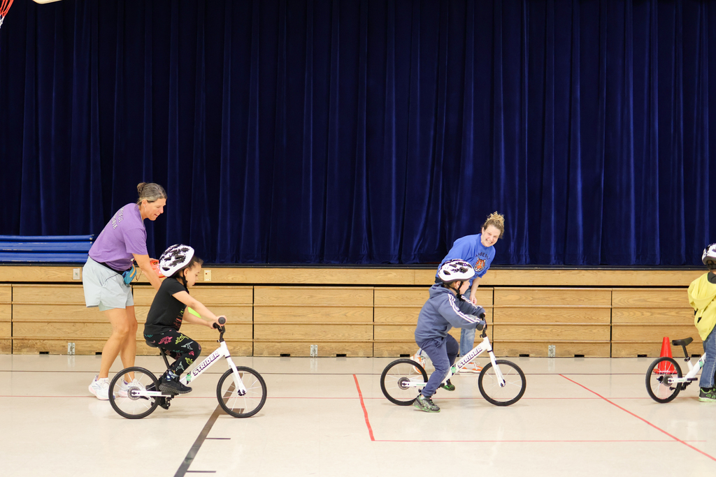 students riding bikes in a gym while teachers help
