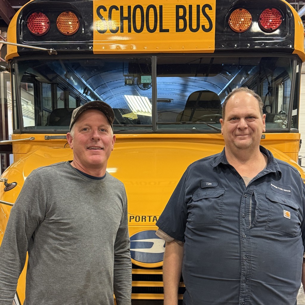 two men pose for a photo in front of a school bus