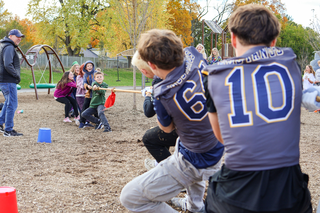 football players play tug-of-war against elementary students outside on a playground