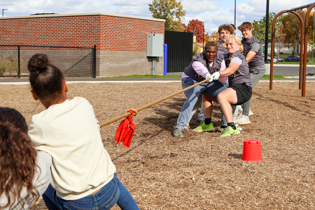 high school football players play tug-of-war against elementary students outside on a playground
