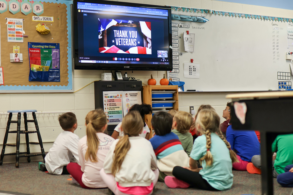 students sitting on the floor of a classroom looking at a tv that says thank you veterans