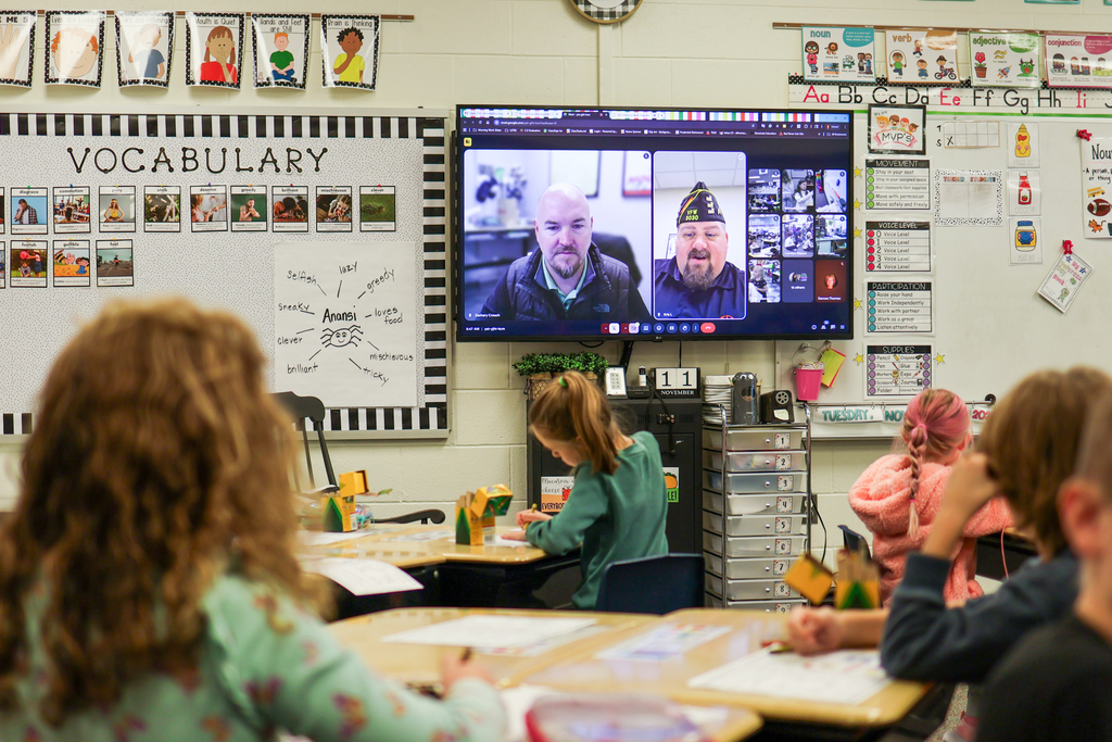 two men speaking on a zoom call projected on a tv in a classroom