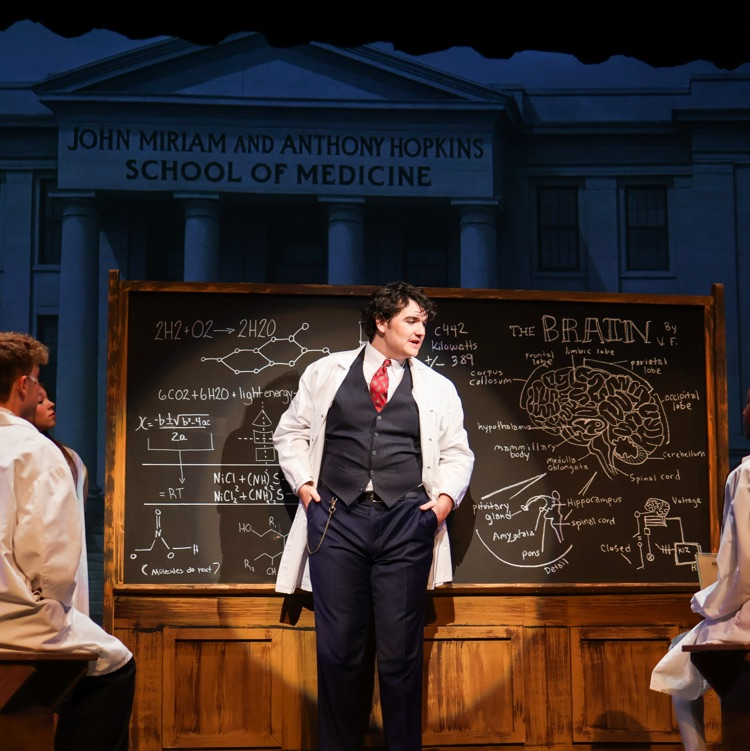 a doctor on stage standing in front of a chalkboard