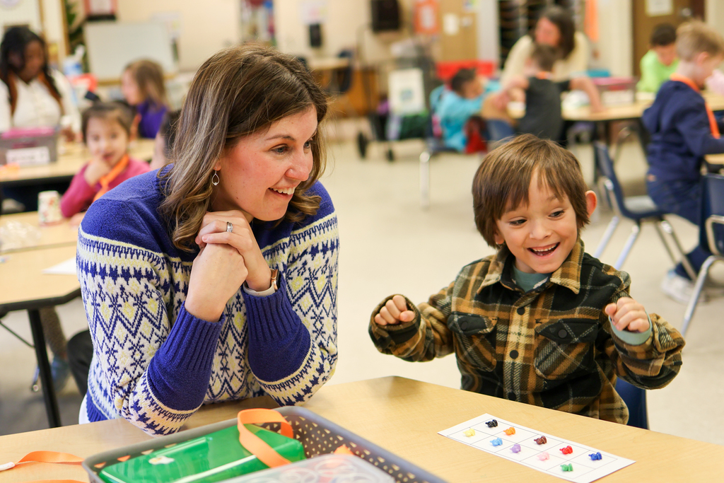 a teacher and student smile together at a desk in a classroom