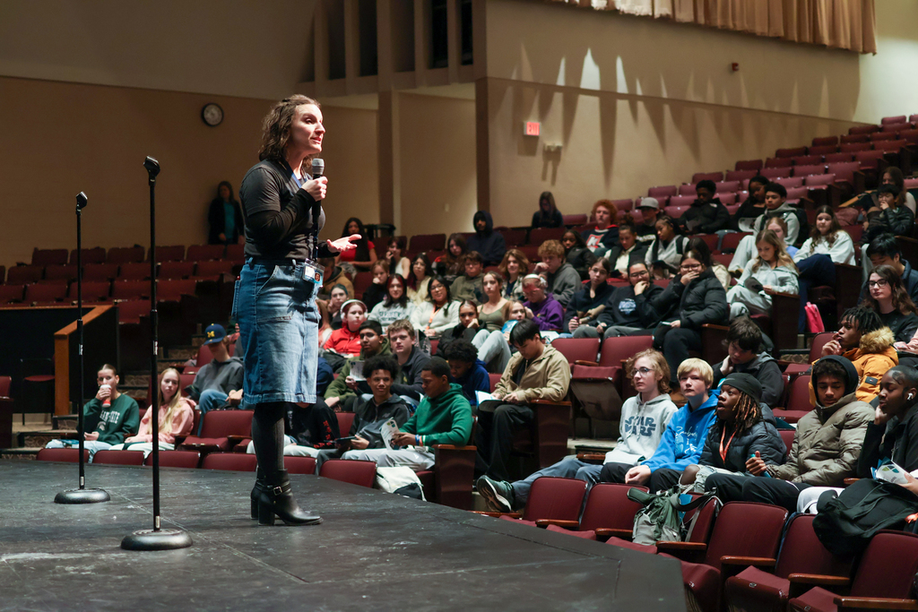 a woman speaks on stage in front of students in an auditorium