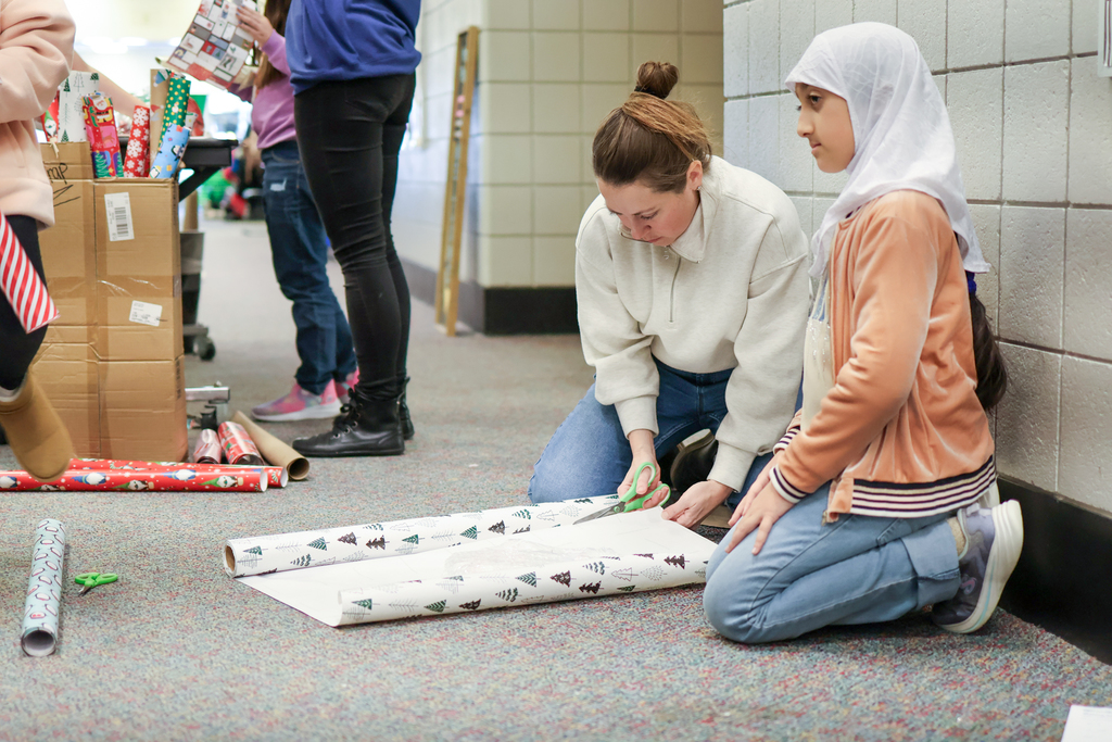 a woman cuts wrapping paper sitting next to a student