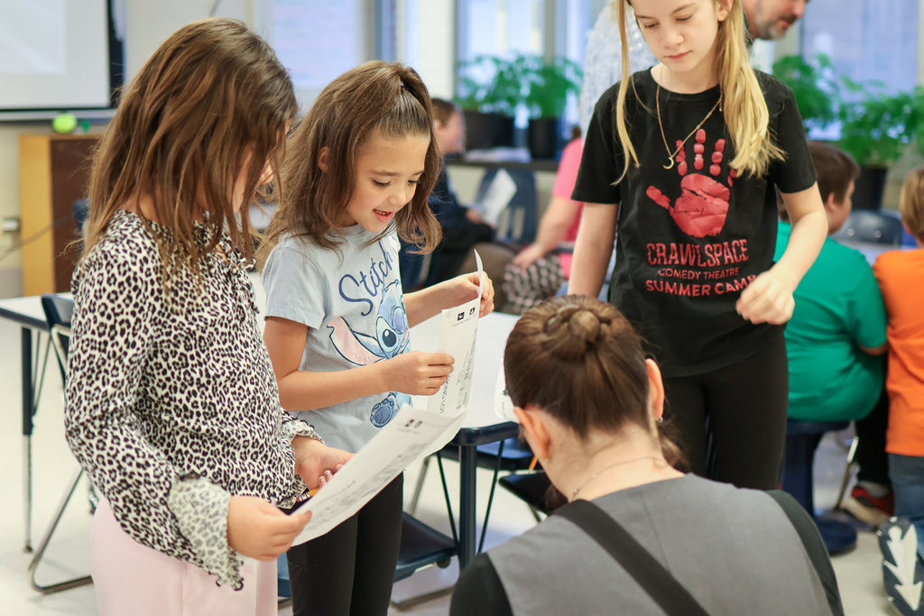 girls holding papers stand and talk to a woman