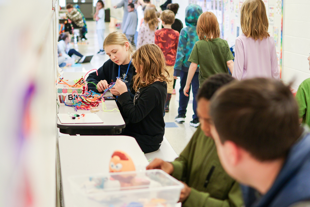high school students sitting next to elementary students at desks in a hallway