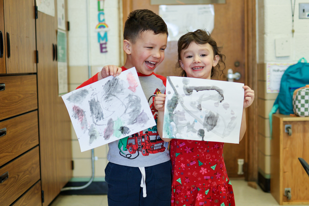 two kids smiling in a classroom holding paintings