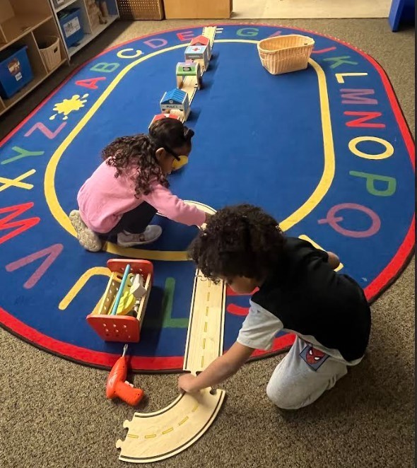 Two preschool students making a long train track on the carpet together.