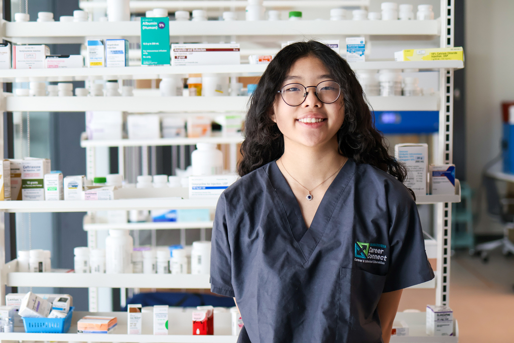 a girl wearing scrubs poses for a photo in front of a shelf of medication