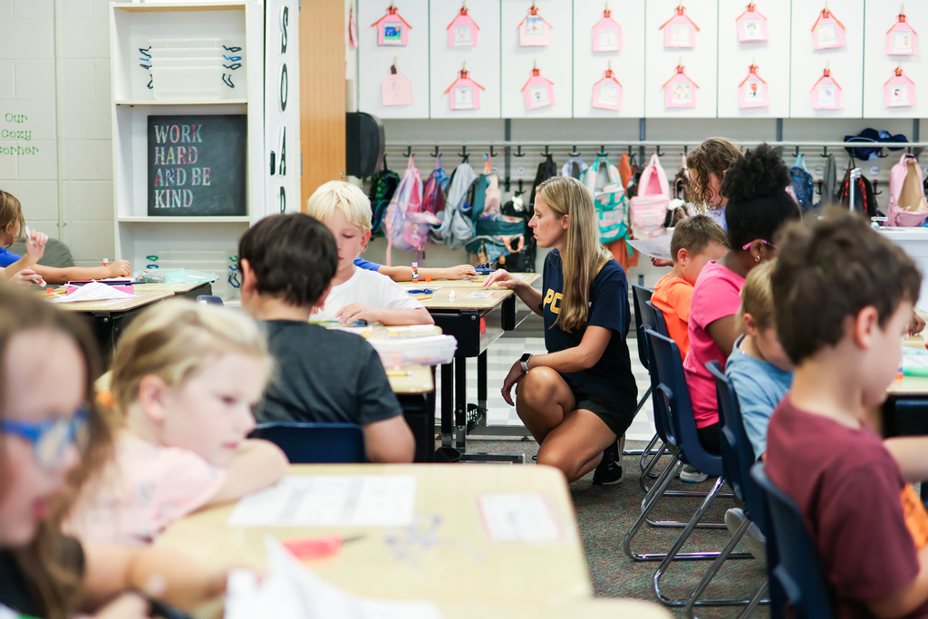 a teacher crouching down beside a student's desk inside a classroom