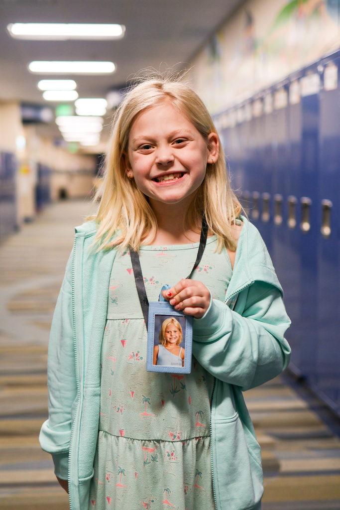 a little girl posing for a photo in a school hallway holding up her "teacher badge." 