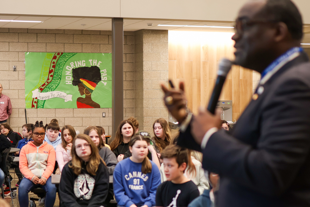 a sign seen behind students gathered in a cafeteria listening to a man speak