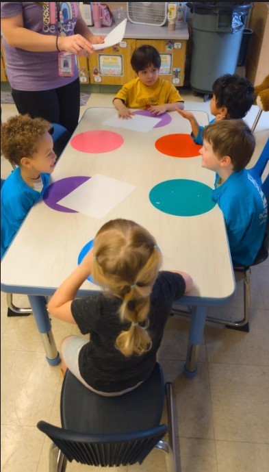 Students getting paintbrushes from their teacher at the table.