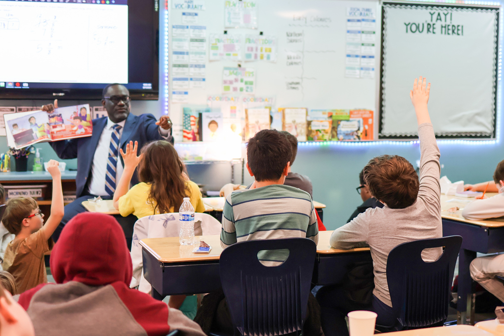 a boy raising his hand in class while a man points at him from the front of the classroom
