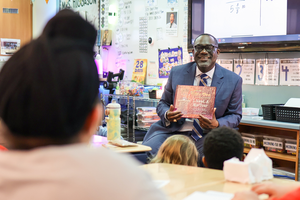 a man holding a book smiles at students sitting in a classroom