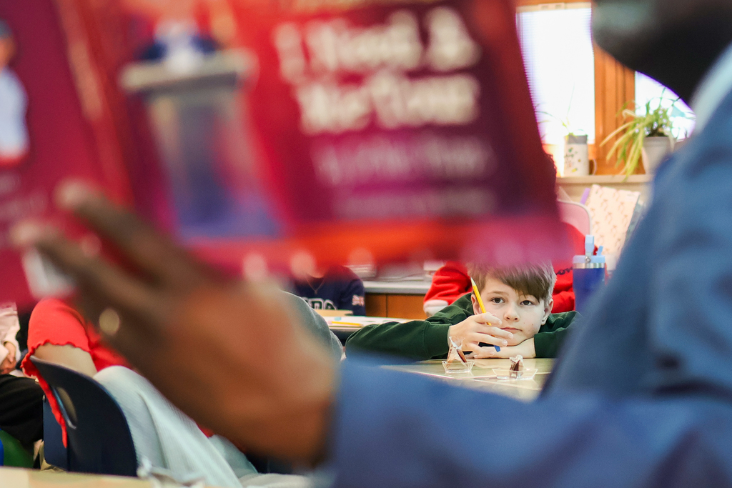 a little boy is seen sitting at his desk through the arms of a man reading a book in a classroom