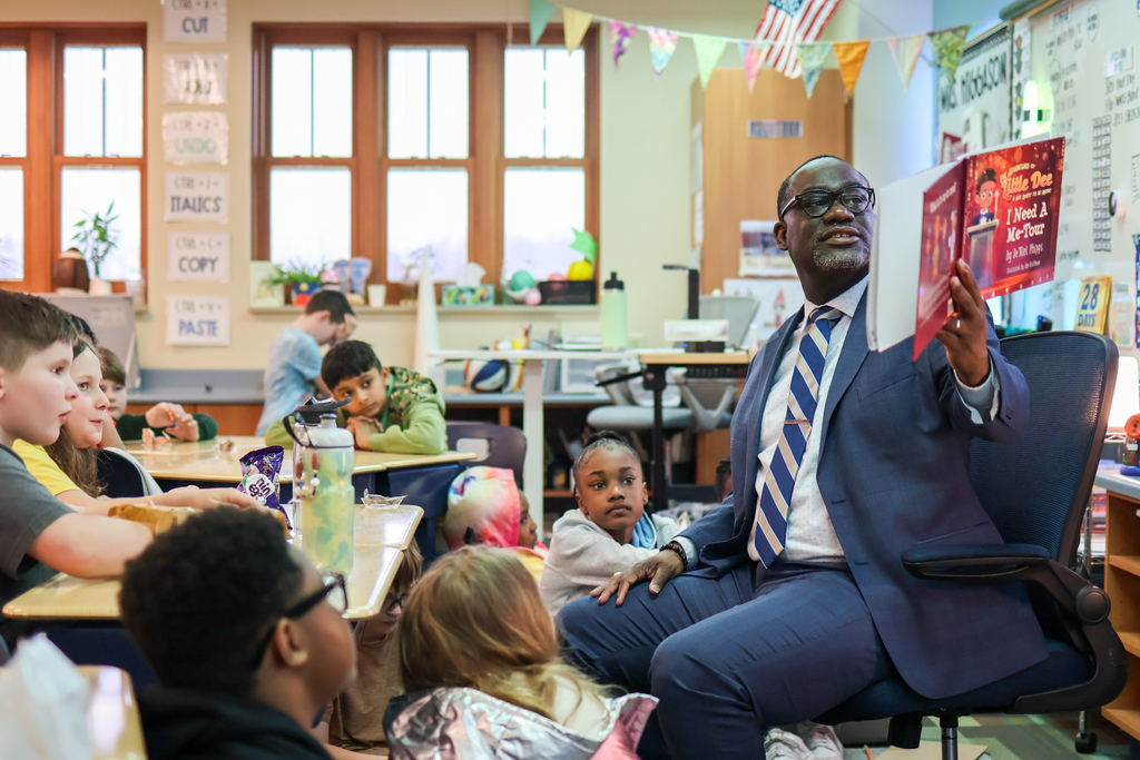 children listen as a man reads a book at the front of a classroom