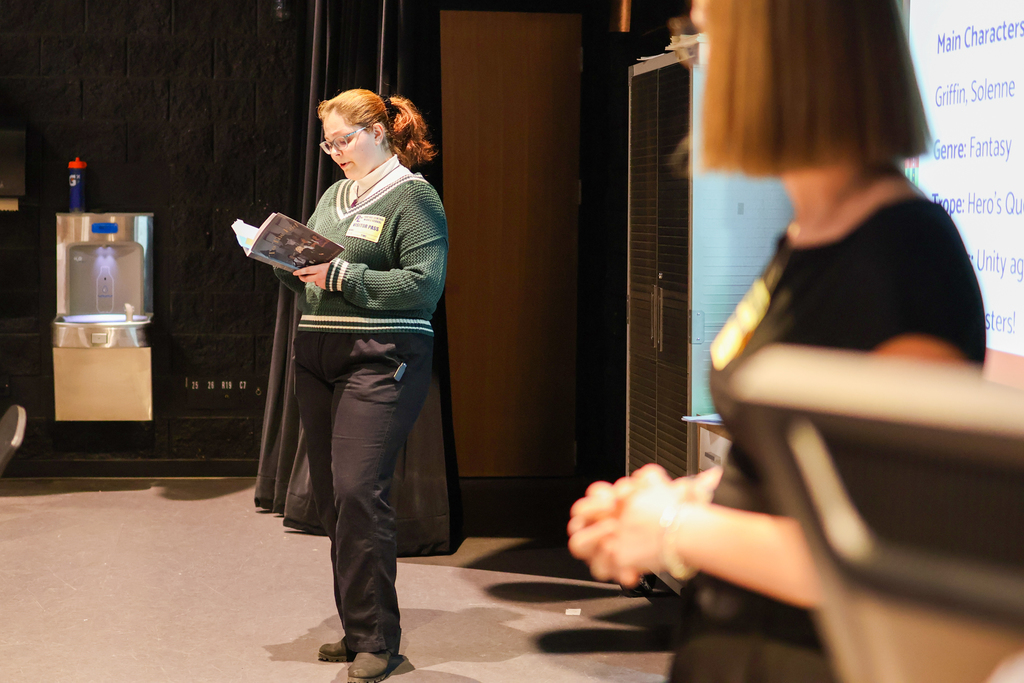 a woman reads from a novel standing at the front of a classroom