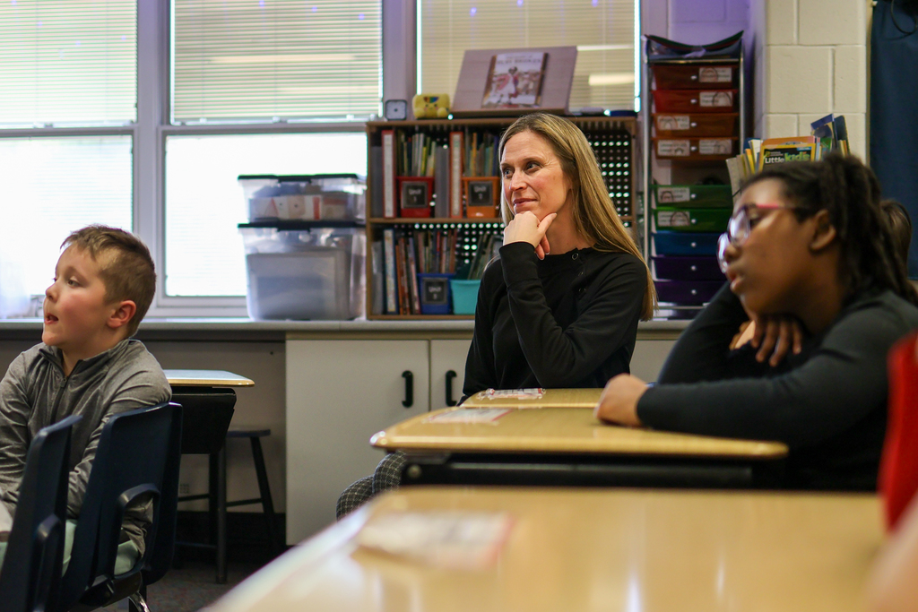a teacher and students sit at desks listening in a classroom