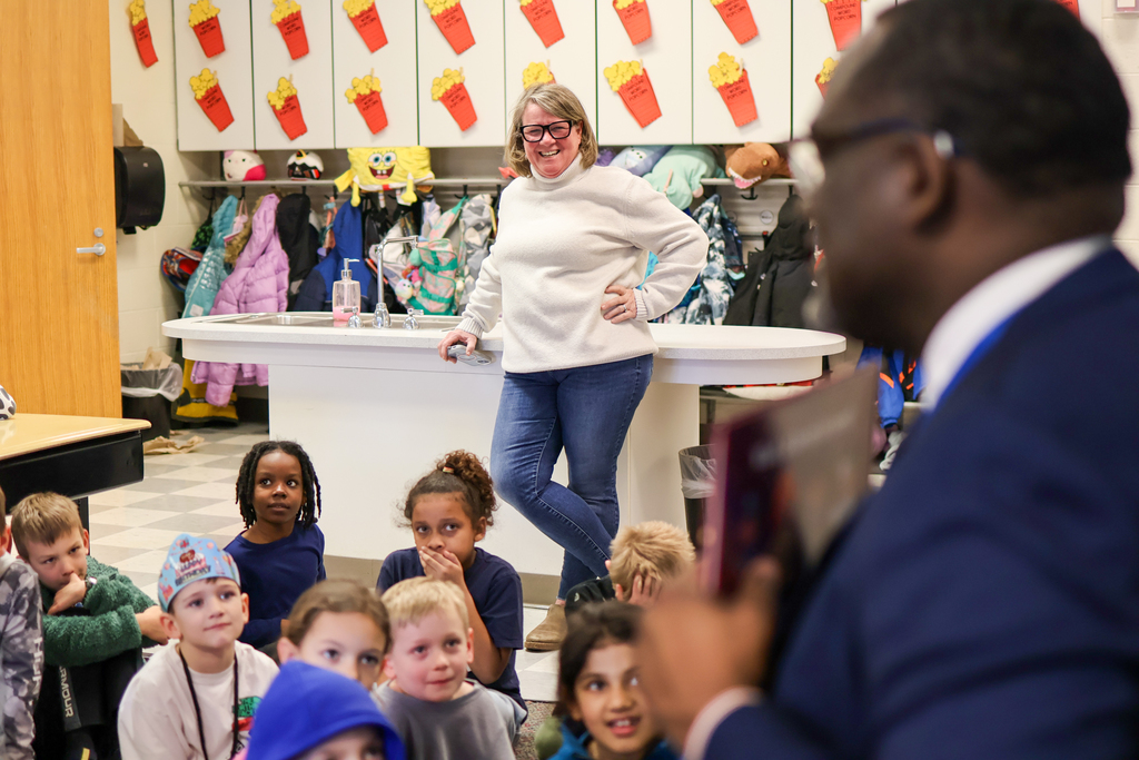 a teacher smiles standing up behind students sitting on the floor in a classorom