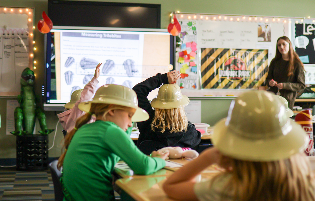 students seen from the back of the classroom raising their hands while a teacher speaks from the front