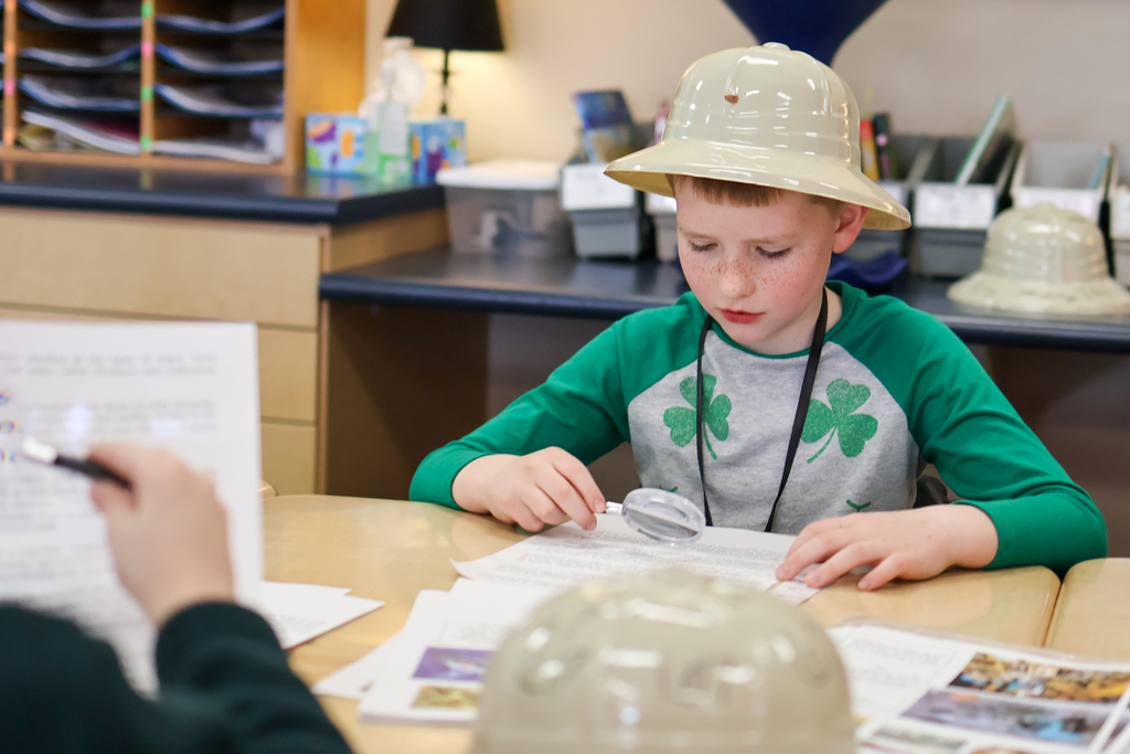 a boy wearing a scientist hat using a magnifying glass to read a paper
