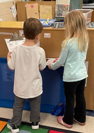A boy and a girl student holding clipboards in their classroom.