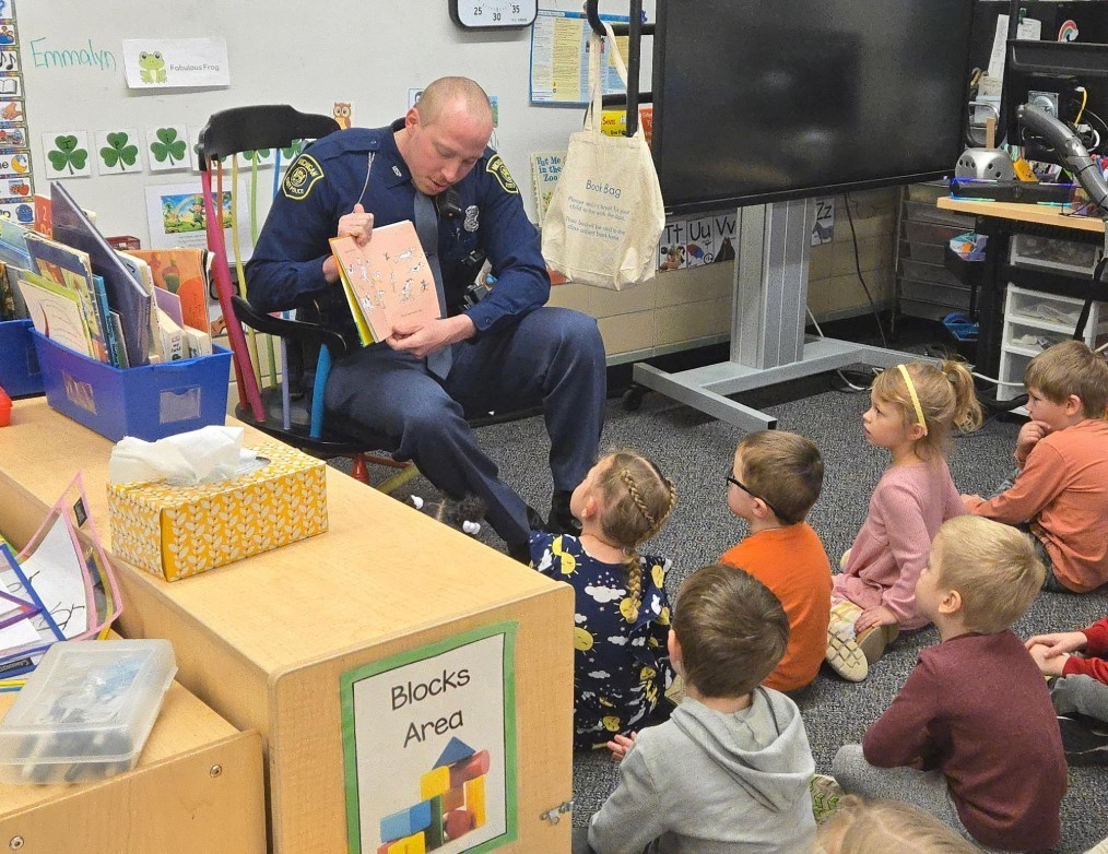 A male state trooper reading a book to a group of four year old children.