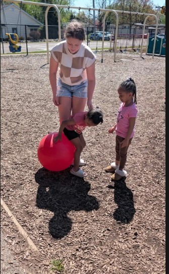 A 8th grade girl helping two female three year old students on a ball.