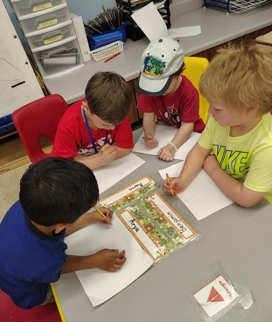 A big kid and three younger boys tracing their names at the table together. 
